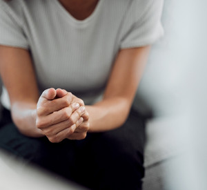 Abgeschnittene Aufnahme: Eine nicht wiederzuerkennende Frau sitzt allein mit gefalteten Händen.