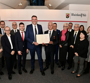 Minister Alexander Schweitzer und Bernd Meurer halten inmitten einer Gruppe die Urkunde in die Kamera 