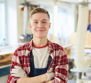 Ein Zimmerei-Student steht mit Arbeitshandschuhen in einer Werkstatt und schaut frontal in die Kamera.
